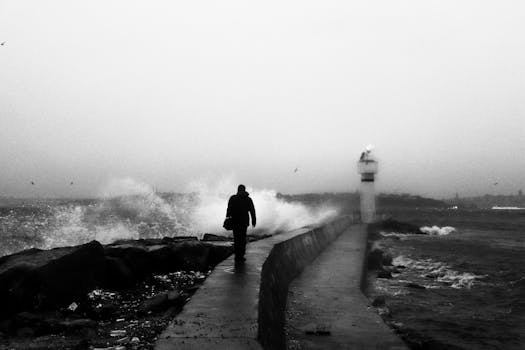 A lone silhouette walking on a breakwater with crashing waves and lighthouse in monochrome.