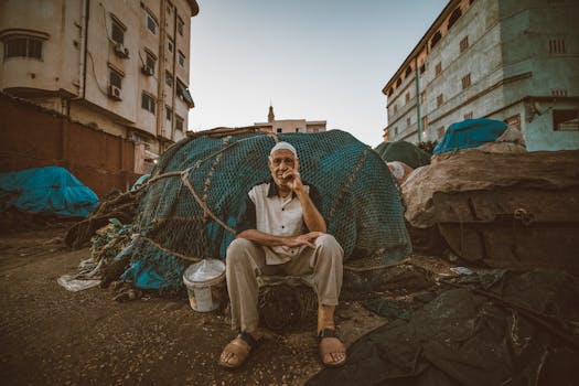 An elderly fisherman sits surrounded by fishing nets in Ras El-Bar, Egypt, at dusk, showcasing a scene of local resilience.